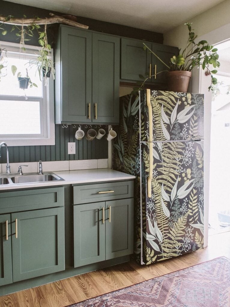 Refrigerator covered in dark botanical wallpaper with tropical leaves in a green kitchen.