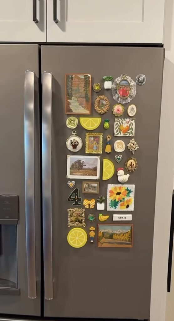 Colorful fridge gallery wall on a stainless steel refrigerator with tiny framed art, photos, flowers, fruit, miniature plants and playful decorative magnets.